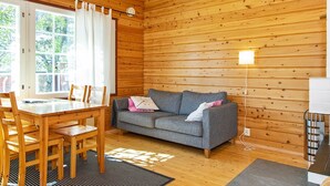 Interior acogedor de una cabaña con techo de madera, un sofá gris, una mesa de comedor de madera con sillas y un amplio ventanal con cortinas.
