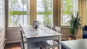 Ein Essbereich mit Tisch und Stühlen, große Fenster mit Blick auf Bäume und Wasser, und ein Topfpflanze.
