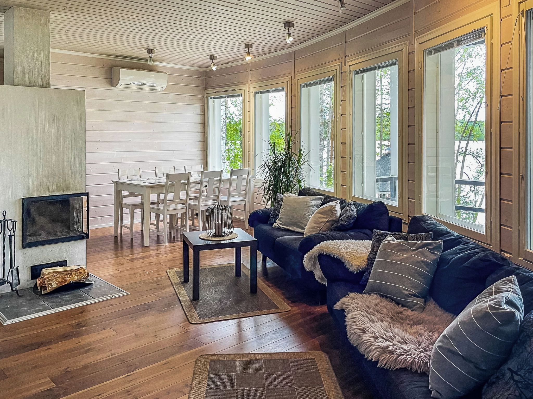 A spacious living room with a dark-colored sofa, a coffee table, and a fireplace. There are multiple windows providing natural light and views of the outdoors.