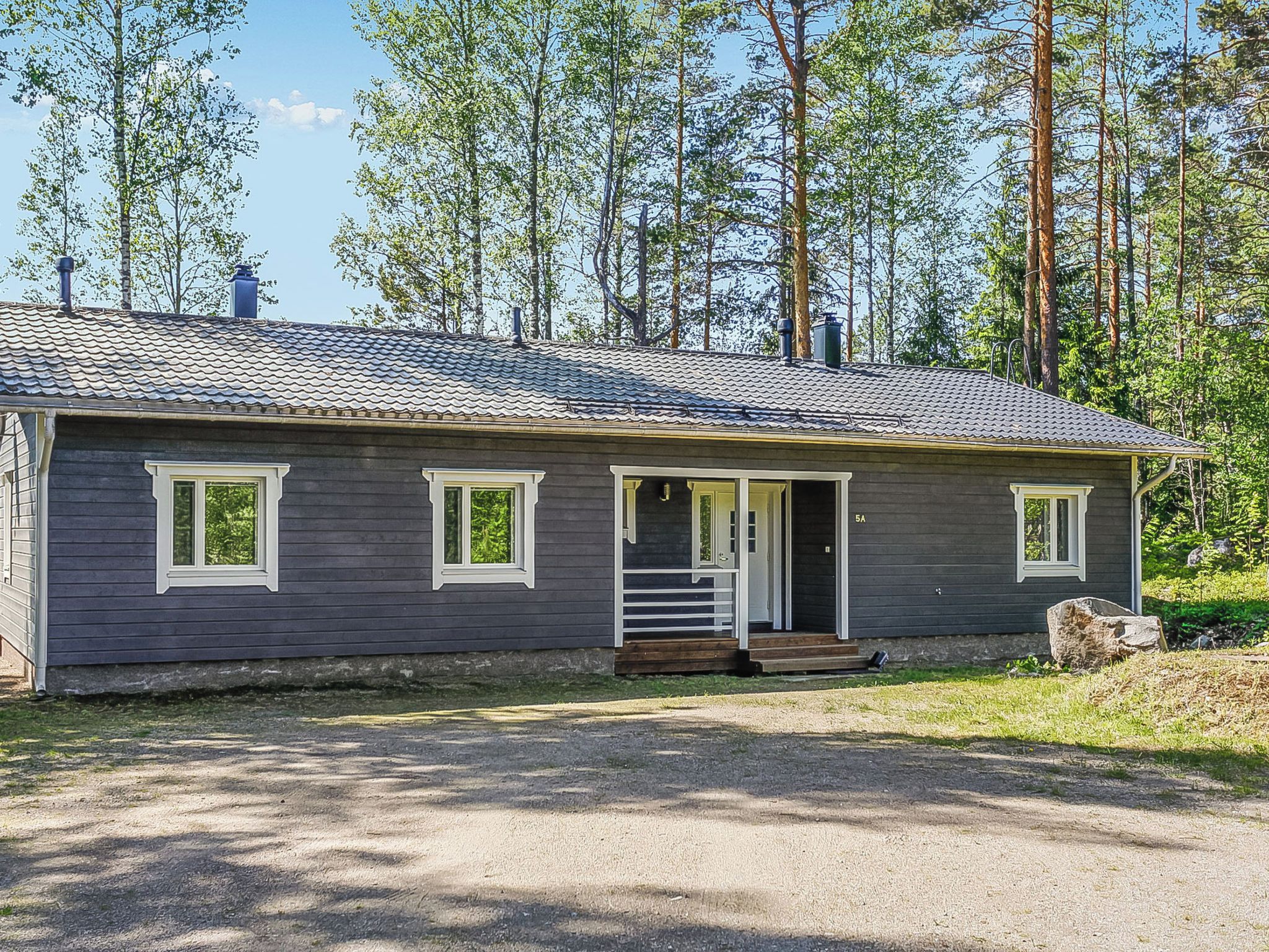 Yhden kerroksen mökki, jossa on etukuisti, savupiippu ja sorapäällysteinen ajotie.