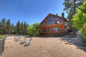 A two-story wooden cabin with a balcony, surrounded by trees and a sandy area with beach chairs. - Lakeview Lodge By Destination Big Bear: Lakefront Luxury with Dock & Views! Hot Tub! Near Village! (Big Bear Lake)