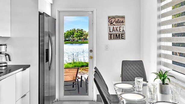 Una cocina moderna con un refrigerador de acero inoxidable, un área de comedor con una mesa dispuesta para dos personas, y vista a un lago a través de una puerta.