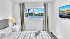 Habitación de hotel con cama, mesita de noche con lámpara, vista a un lago y un cuadro en la pared.