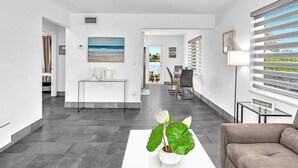 Una sala de estar moderna con una mesa de centro blanca, una planta en maceta y un sofá marrón. Hay un escritorio con una computadora y una silla, y un comedor con mesa y sillas.