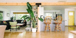 A modern living room with a black sofa, green armchair, and a dining area with two wooden chairs. A large potted plant is in the center. - Lakeside PEC: Beautiful Waterfront & Patio (Wellington)