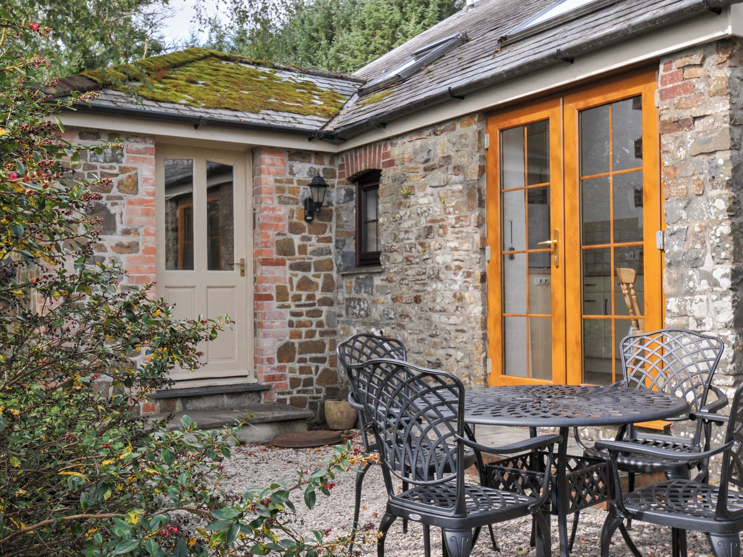 A stone building with a wooden door and windows, outdoor seating with metal chairs and a table, and a gravel ground.