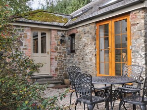 A stone building with a wooden door and windows, outdoor seating with metal chairs and a table, and a gravel ground. - Shipload Cottage (Bideford)