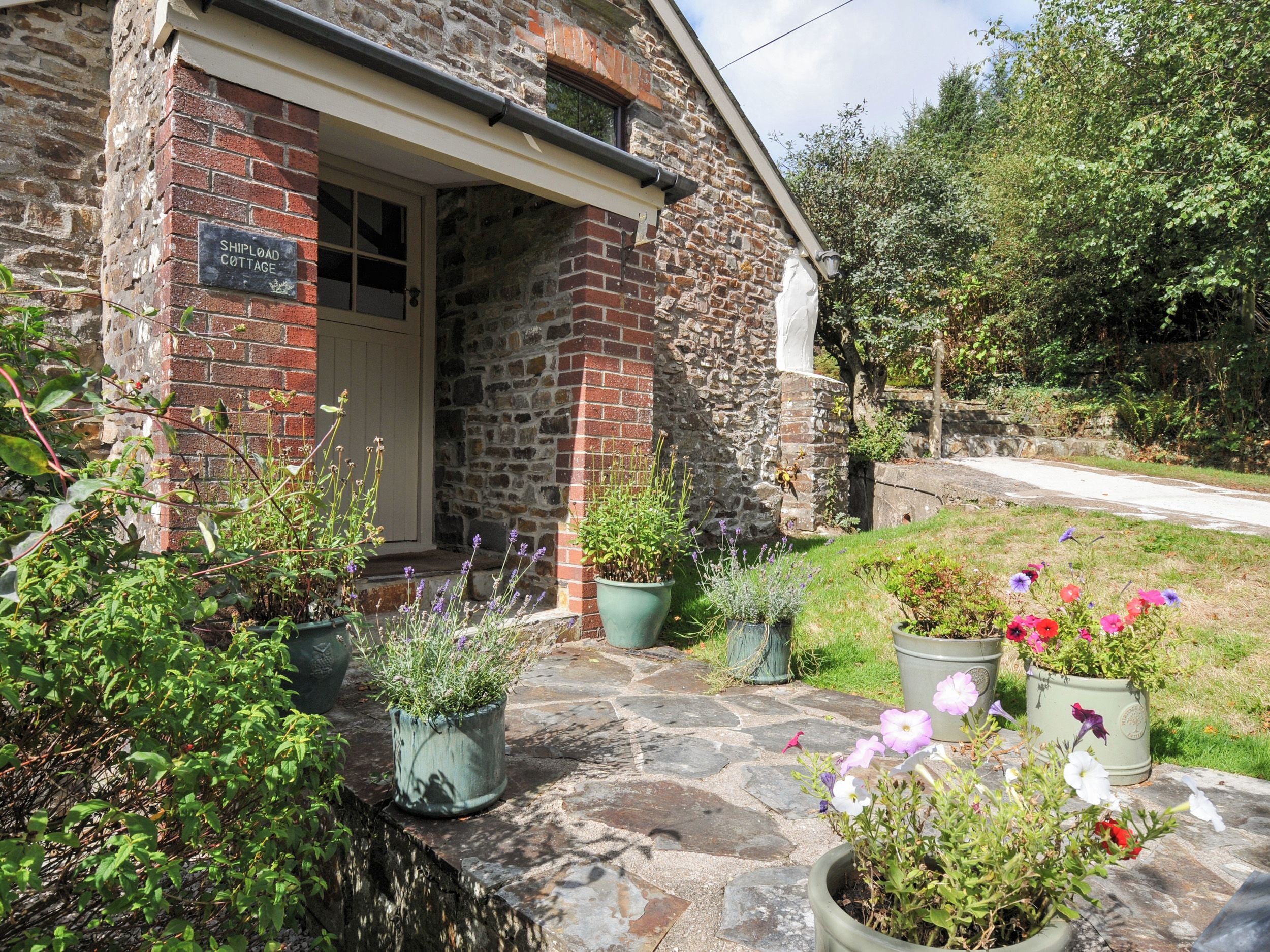 A stone cottage with a sign reading 'Shipload Cottage,' surrounded by potted plants and flowers.
