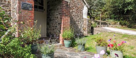 A stone cottage with a sign reading 'Shipload Cottage,' surrounded by potted plants and flowers.