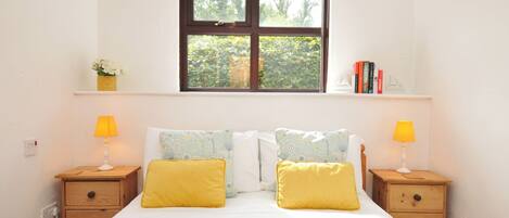 A bedroom with a bed, two bedside tables, a window with a view, and a shelf with books and a plant.