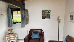 A cozy living room with a brown leather armchair, a wooden side table with a vase of flowers, and a blue cushion. - Shipload Cottage (Bideford)