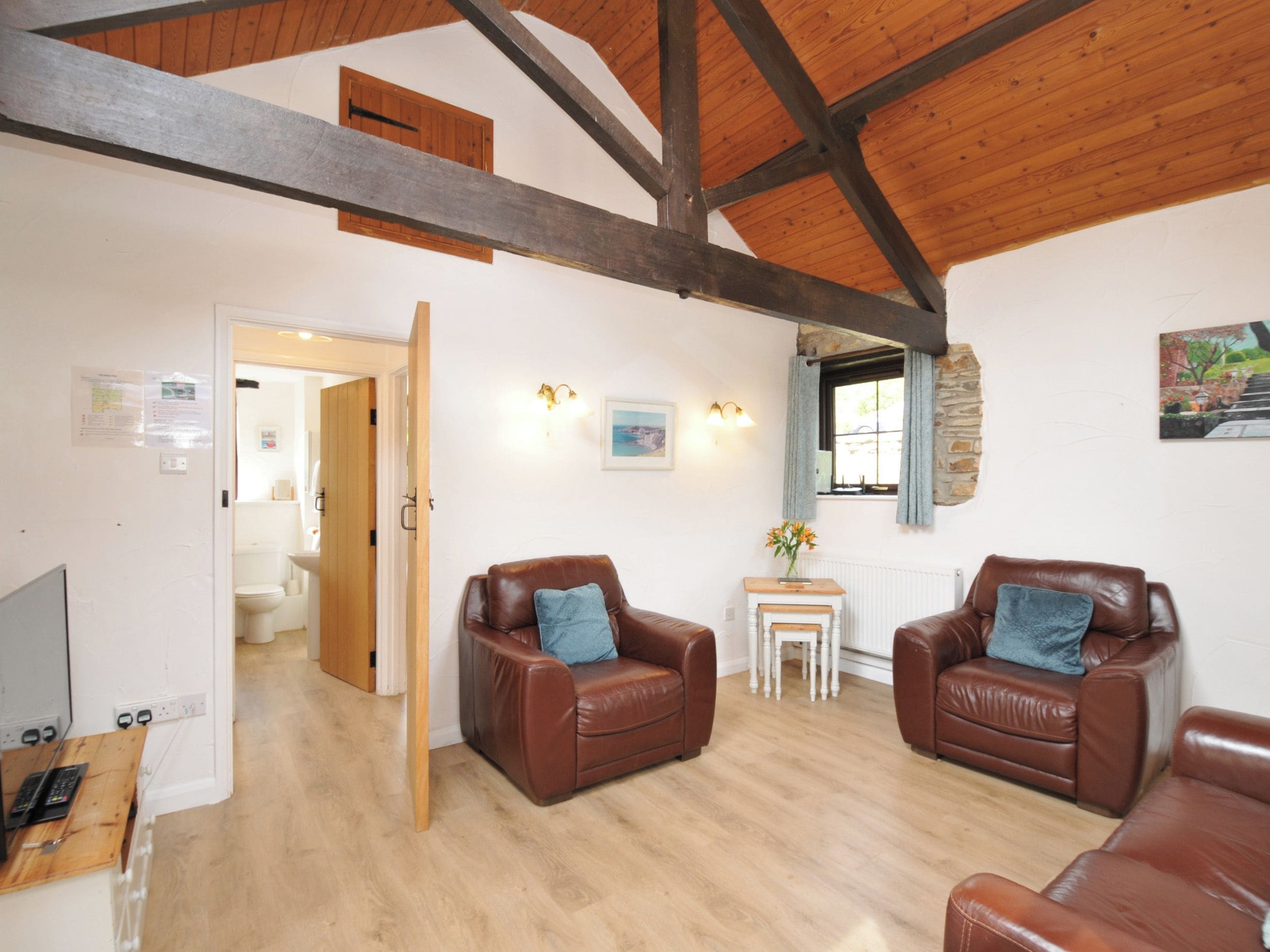 A spacious room with a high ceiling, wooden beams, two leather armchairs, a small white table, and a view of a bathroom through an open door.