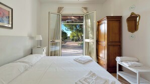 A hotel room with a large bed, a wardrobe, a bedside table with a lamp, and a view through an open door to a garden. - VILLA TWO SORDS, with swimming pool, olive grove, orchard, tropical garden! (Ugento)
