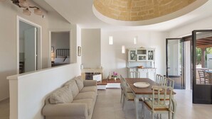A spacious living area with a sofa, dining table, and chairs. A kitchen with cabinets and a dining table is visible in the background. - VILLA TWO SORDS, with swimming pool, olive grove, orchard, tropical garden! (Ugento)