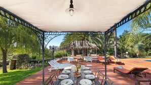An outdoor dining area with a covered table set for a meal, surrounded by lounge chairs and a pool in the background. - VILLA TWO SORDS, with swimming pool, olive grove, orchard, tropical garden! (Ugento)