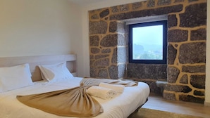 Ein Schlafzimmer mit Steinwand, großem Fenster, Bett mit weißer Bettwäsche und einer gewebten Decke.