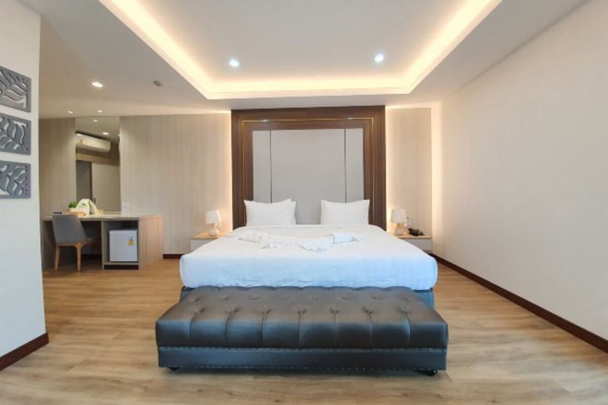 A modern hotel room with a large bed, a bedside table, a desk with a chair, and a small kitchenette.