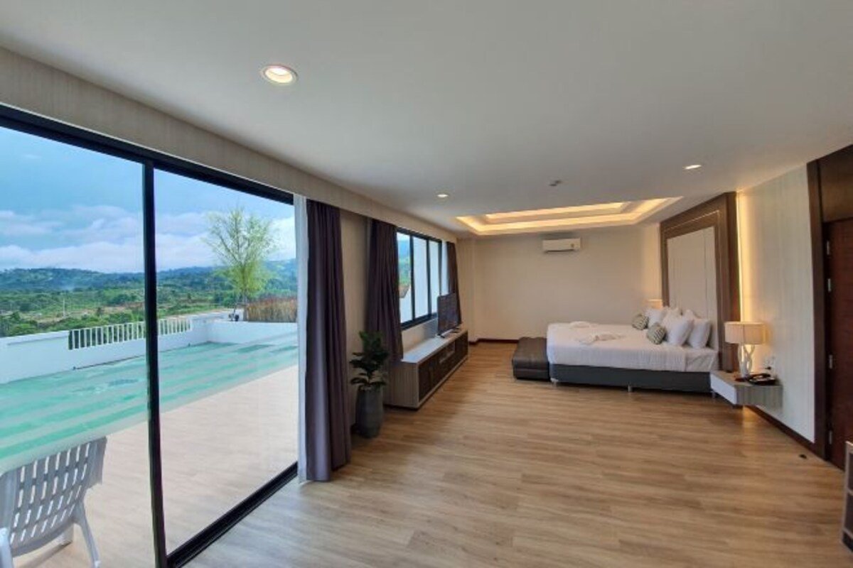 A modern hotel room with a large bed, a TV, a bedside table, and a view of a green landscape through large windows.