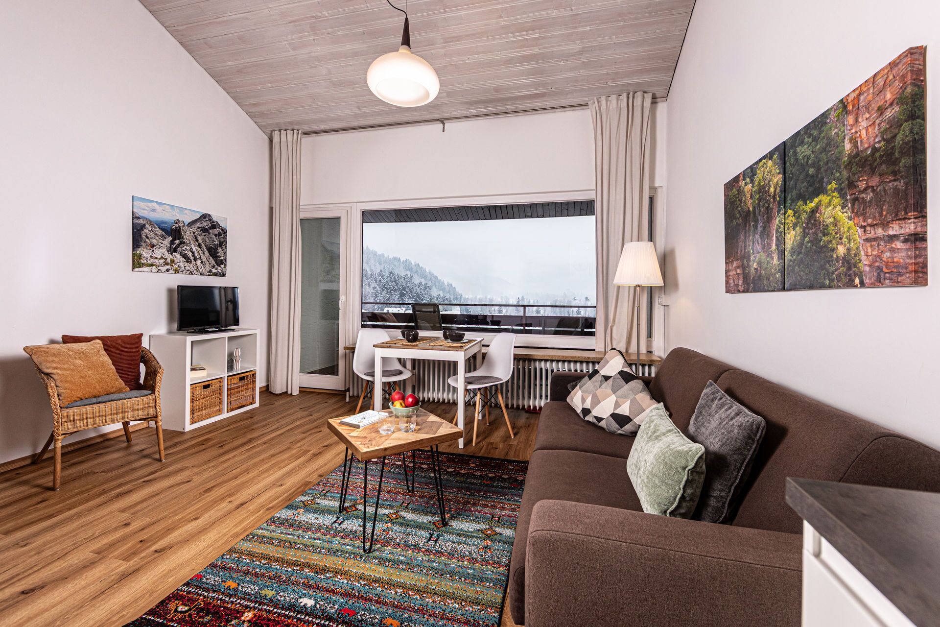 Ein modernes Wohnzimmer mit einer Couch, einem Fernseher, einem Esstisch und Blick auf eine verschneite Landschaft durch ein großes Fenster.