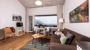 Ein modernes Wohnzimmer mit einer Couch, einem Fernseher, einem Esstisch und Blick auf eine verschneite Landschaft durch ein großes Fenster.