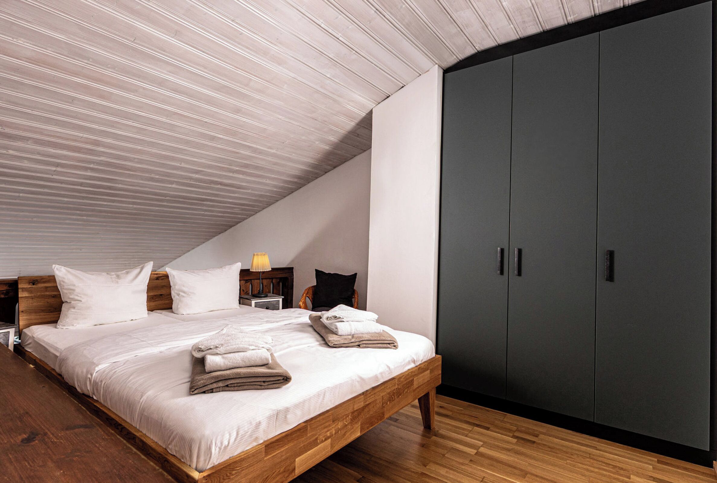 Ein Schlafzimmer mit schrägem Dach, einem hölzernen Bett, einem dunklen Kleiderschrank und einem Nachttisch mit Lampe.