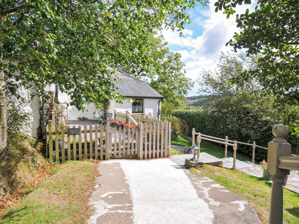 A white cottage with a wooden fence, a gravel path leading to the entrance, and a view of the countryside. - Windbury Cottage (Bideford)