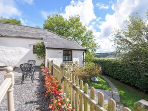 A white cottage with a stone roof, a wooden fence, and a gravel path leading to a seating area with chairs. - Windbury Cottage (Bideford)