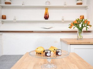 A kitchen with a wooden table displaying muffins and jars, a white shelf with decorative items, and a vase with flowers.