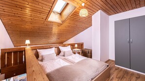 Une chambre avec un plafond en pente, des poutres en bois, un lit avec une literie blanche, une table de chevet avec une lampe, une armoire grise et une fenêtre laissant entrer la lumière naturelle.