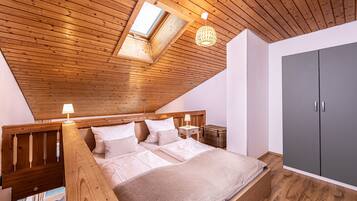 Ein Schlafzimmer mit geneigter Decke, Holzbalken, einem Bett mit weißer Bettwäsche, einem Nachttisch mit Lampe, einem grauen Kleiderschrank und einem Fenster mit Tageslicht.