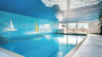 Ein hallenbad mit blau gefliesten Wänden, einem Segelboot-Design, einer klaren Decke und Blick ins Freie durch große Fenster.