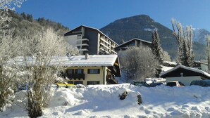 Un bâtiment enneigé, doté de nombreux balcons, entouré d’arbres chargés de neige et avec une montagne en arrière-plan.