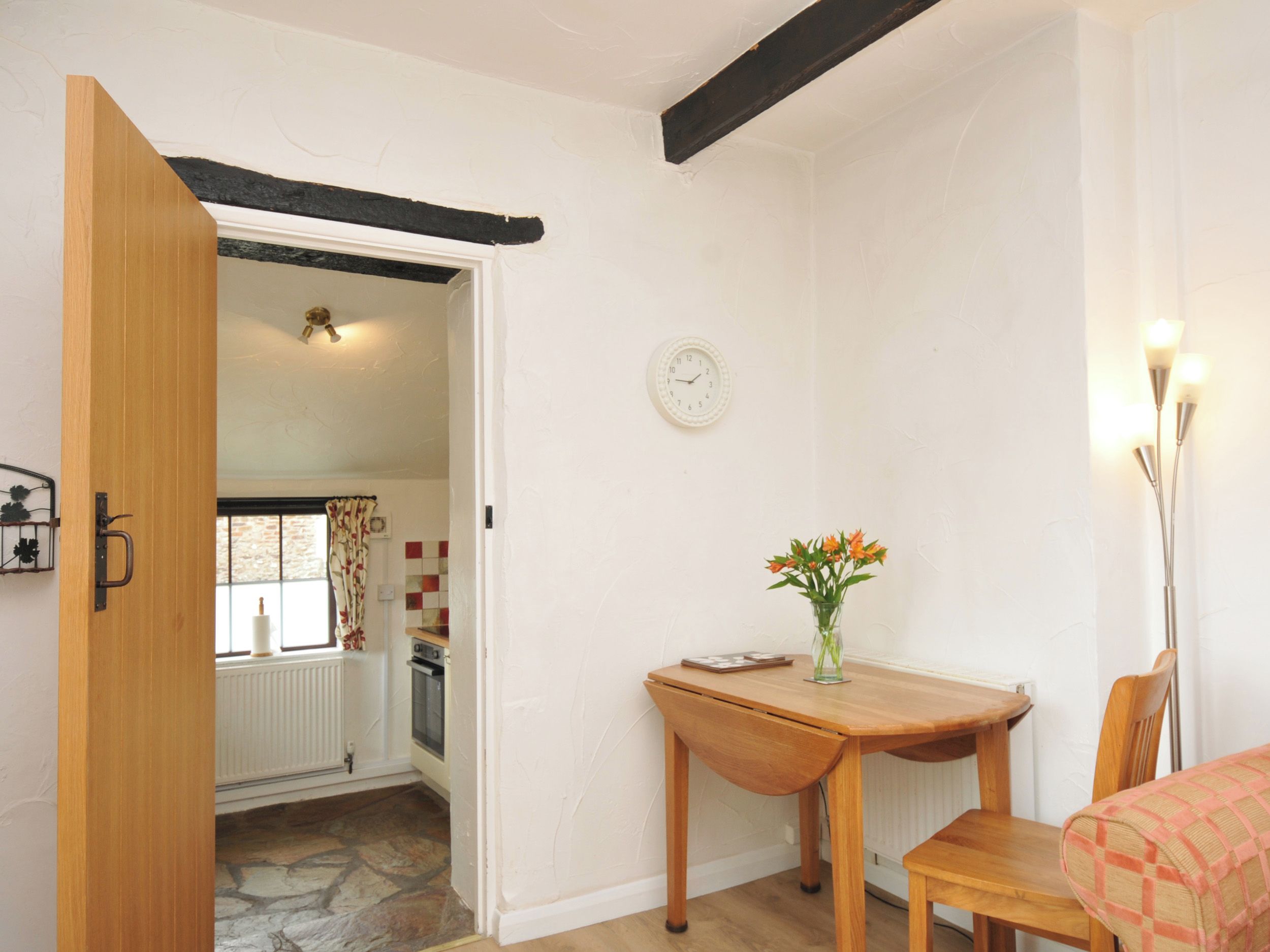 A room with a wooden door, a table with a vase of flowers, a clock on the wall, and a radiator.