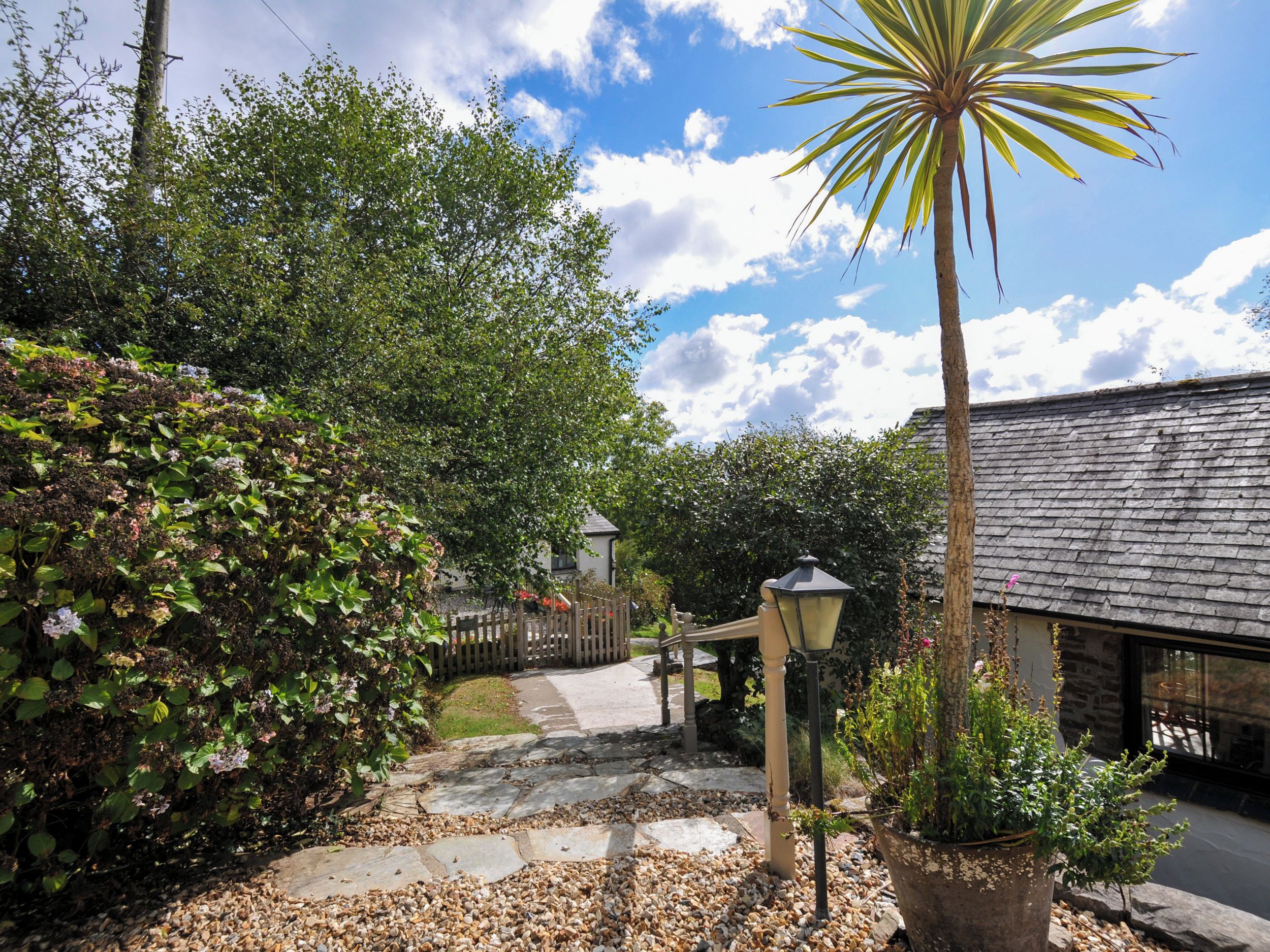 A garden pathway leading to a building with a stone roof, surrounded by lush greenery and a wooden fence.
