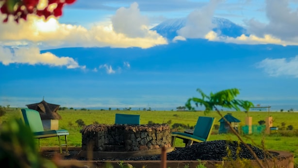 Ein ruhiger Sitzbereich im Freien mit Steinkaminkuppeln und grünen Stühlen, mit Blick auf ein weitläufiges Grasfeld und ferne Bergketten unter blauem Himmel.