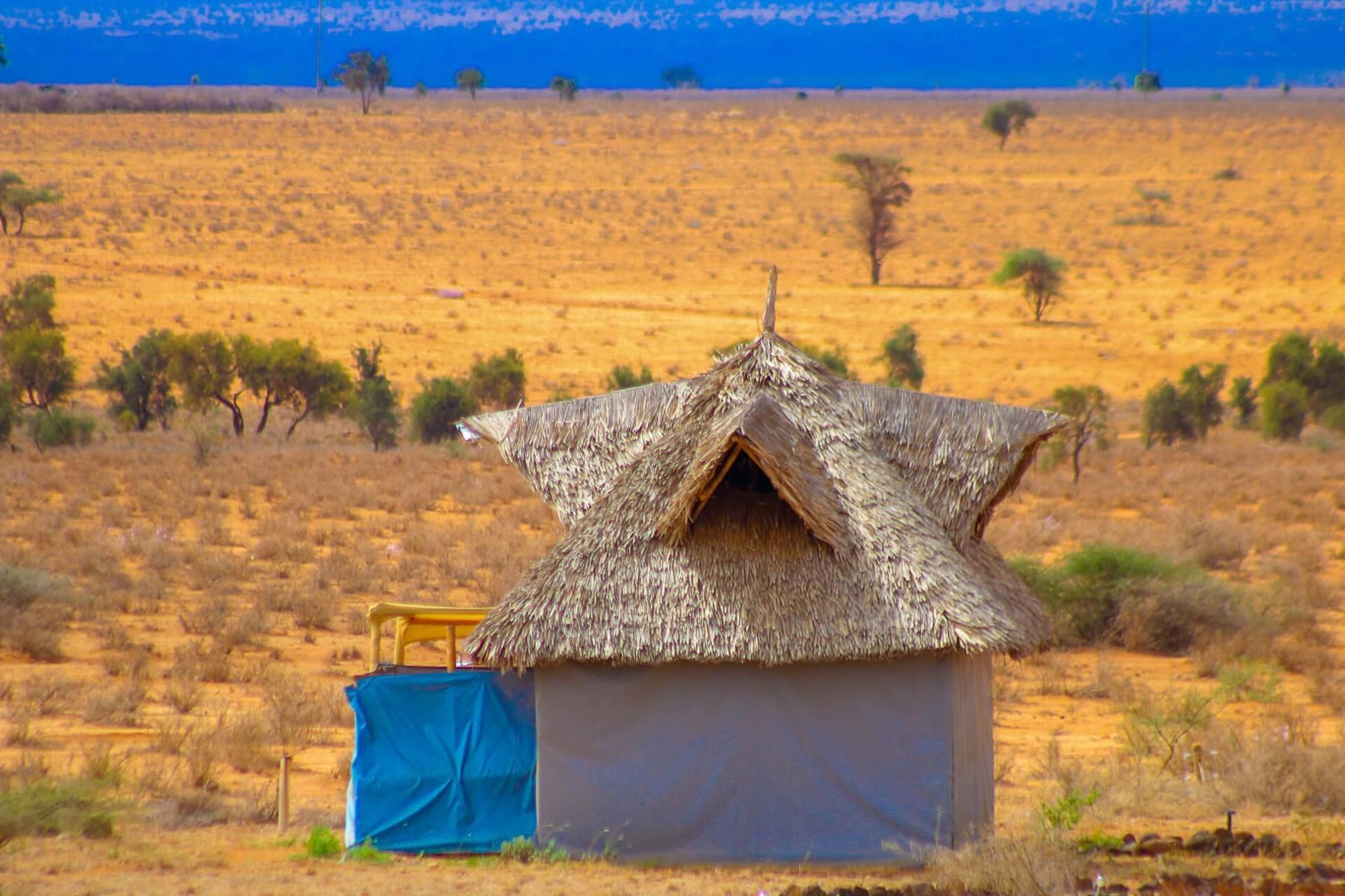 Una pequeña cabaña con techo de paja y un lomo azul cercano, situada en un paisaje extenso y abierto con vegetación escasa.