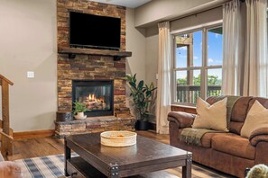 Una sala de estar con chimenea de piedra, un televisor de pantalla plana montado encima, un sofá de cuero marrón y un gran ventanal con cortinas.