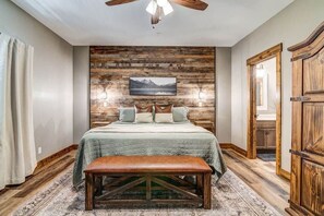 A bedroom with a wooden wall panel, a bed with pillows, a bench, and a ceiling fan.