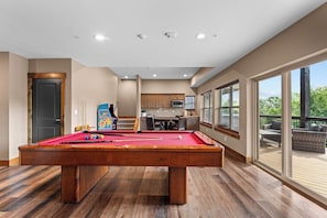 A pool table in a room with a kitchen, dining area, and large windows.