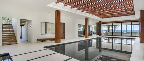 A spacious indoor area with a reflective floor, wooden ceiling beams, and large windows offering a view of the sea.