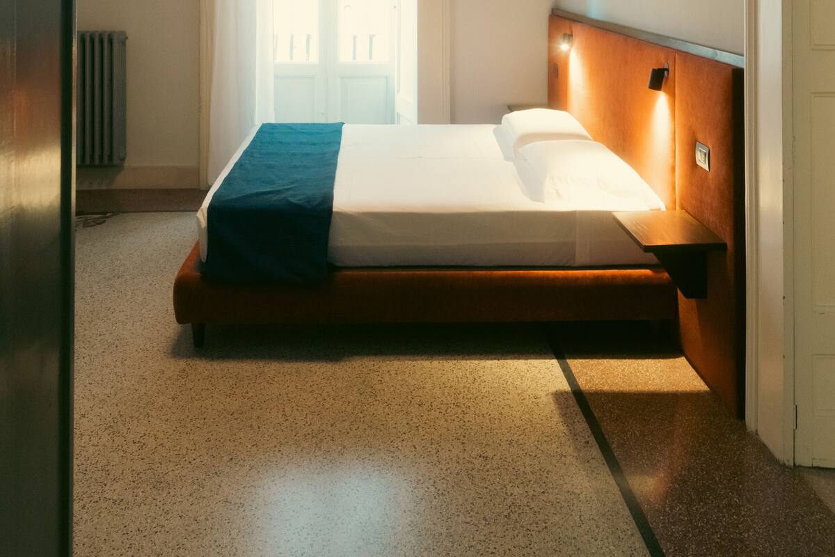 A bedroom with a bed, a radiator, a window with curtains, and a wall-mounted light fixture.