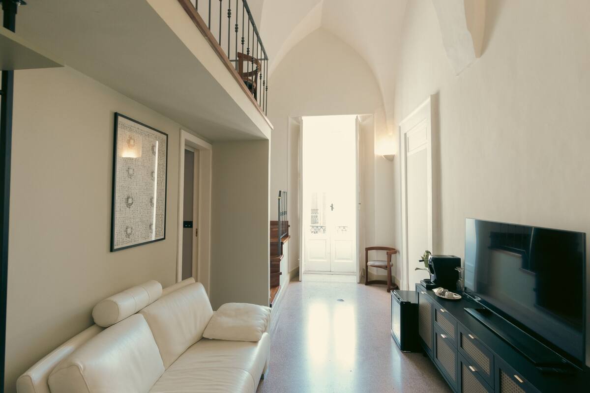 A spacious living area with a high ceiling, a white sofa, a flat-screen TV, and a staircase leading to an upper level.
