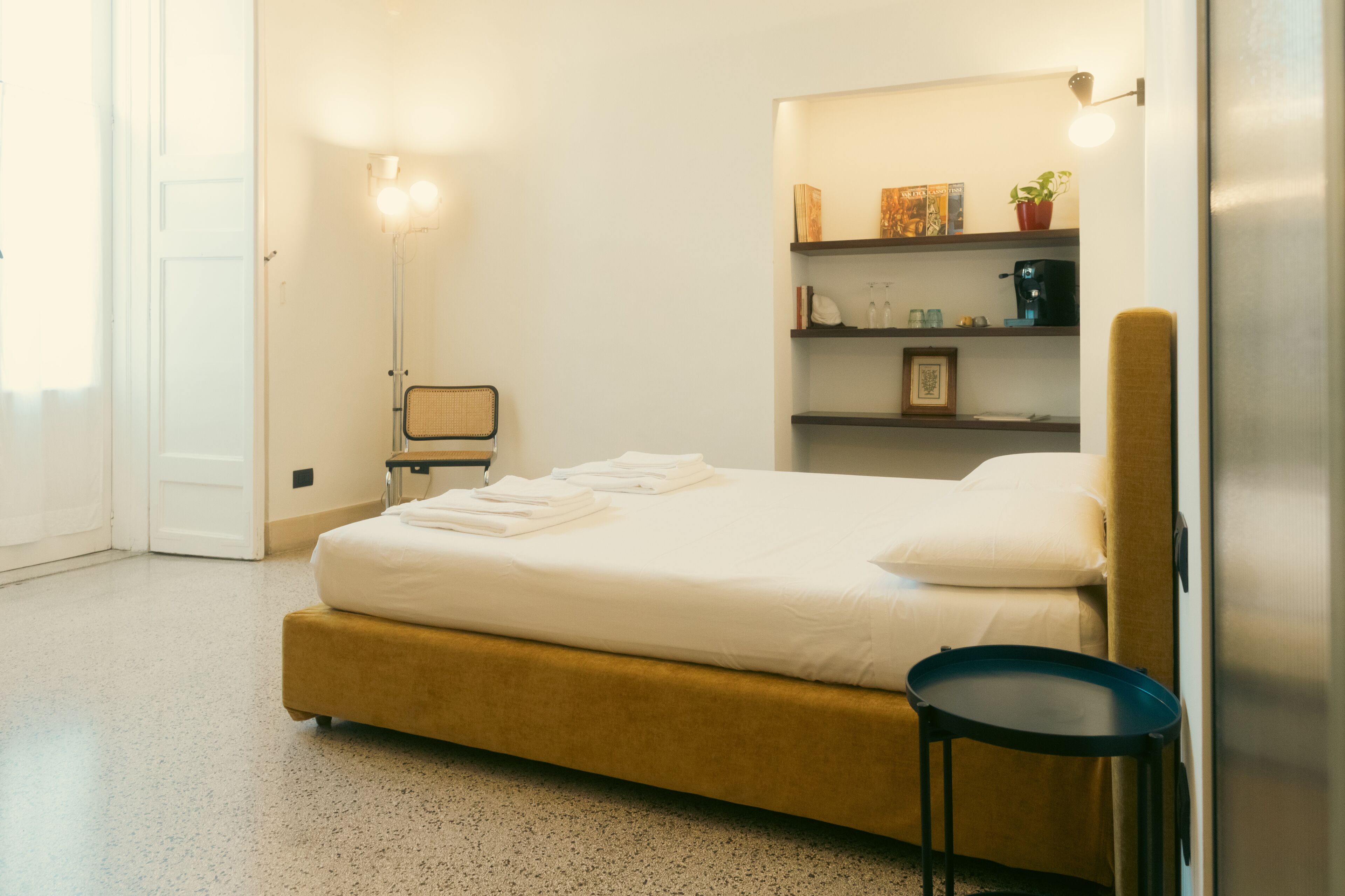 Ein Schlafzimmer mit einem Bett, einem Nachttisch, Regalen mit Büchern und einer Pflanze sowie einer Tür.