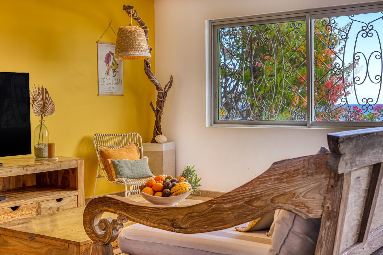 Een gezellige woonkamer met een houten salontafel waarop fruit staat, een rieten fauteuil met kussens, een televisie op een houten dressoir, en een raam met uitzicht op bomen en bloemen.