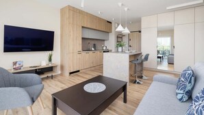 Un salon moderne avec une table basse de couleur sombre, des canapés gris et un coin cuisine doté de meubles en bois et d’un grand téléviseur.