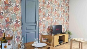 Un comedor con una mesa de madera preparada para dos personas, un televisor sobre un mueble bajo y una puerta con papel tapiz floral.
