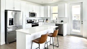 A modern kitchen with white cabinetry, stainless steel appliances, and a central island with bar stools. - Top Floor Luxury Condo (Midvale)
