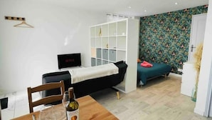 Une pièce moderne et bien éclairée, avec une table à manger en bois dressée pour deux personnes, un lit recouvert d’une courtepointe verte, une armoire et un papier peint à motifs.