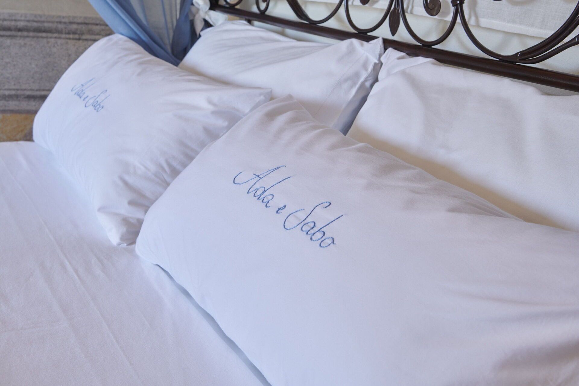 A bed with white linens and pillows, one of which has "Ada e Jabo" embroidered on it.