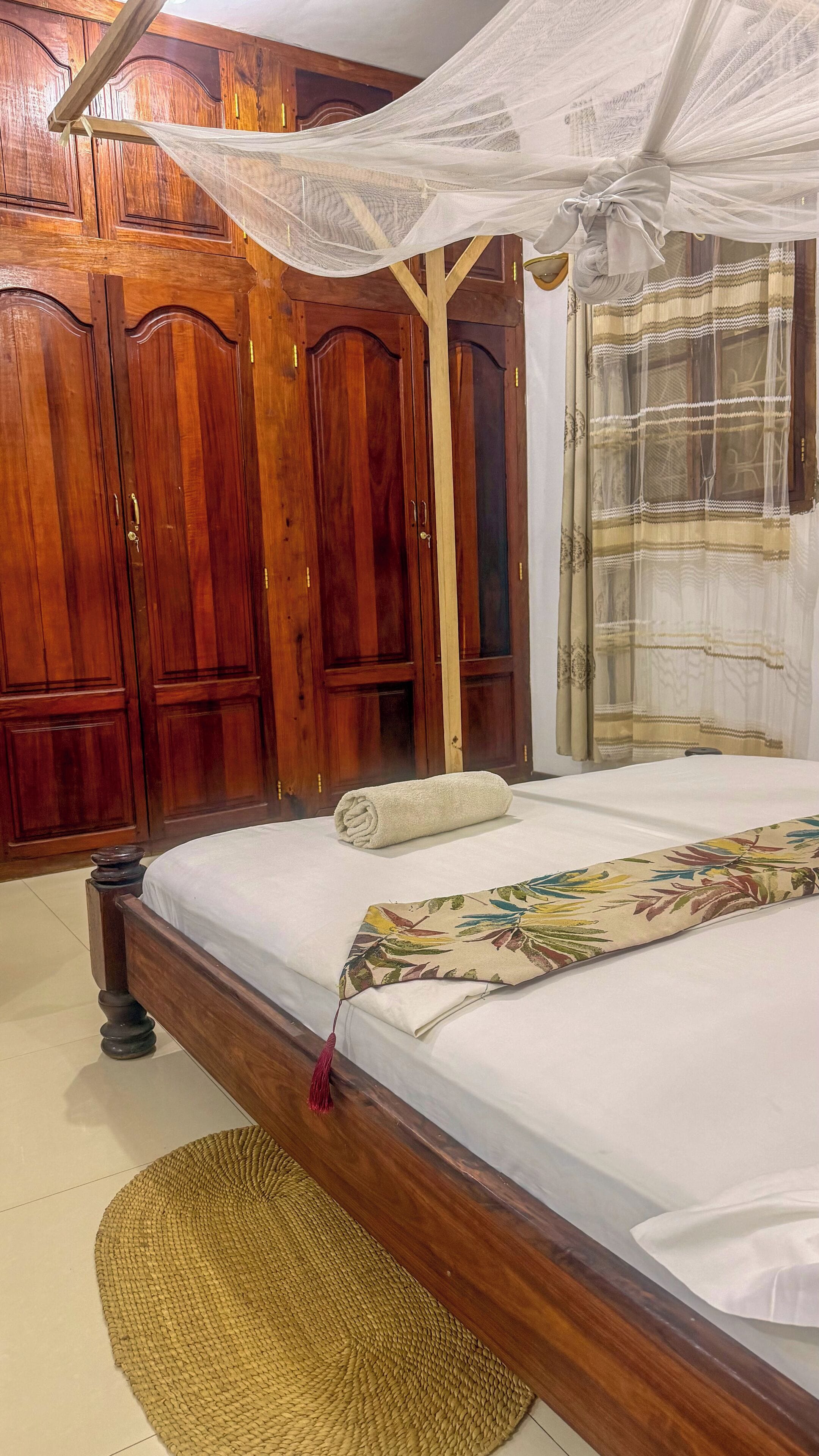 A bedroom with a wooden bed, a mosquito net, a rolled towel, and a woven mat.